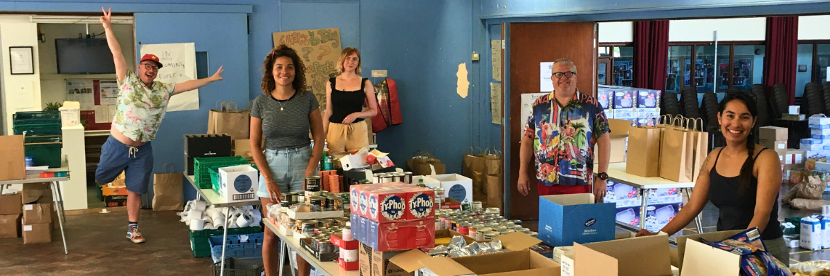 The volunteering team take a break from packing deliveries at the food hub to smile at the camera, surrounded by packages of food.