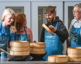 4 people wearing aprons at a kitchen worksurface. one holding the lid to a bamboo stemaer. bamboo steamers on worksurface