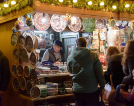 People browsing a festive market stall selling colorful ceramic plates and bowls, with warm lights and greenery decorating the wooden stall at night.